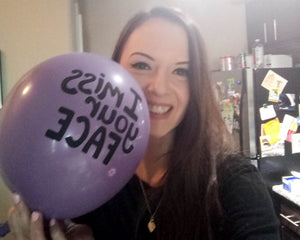 Woman smiling and taking a selfie with a purple balloon by her face with the words "I miss your facce"
