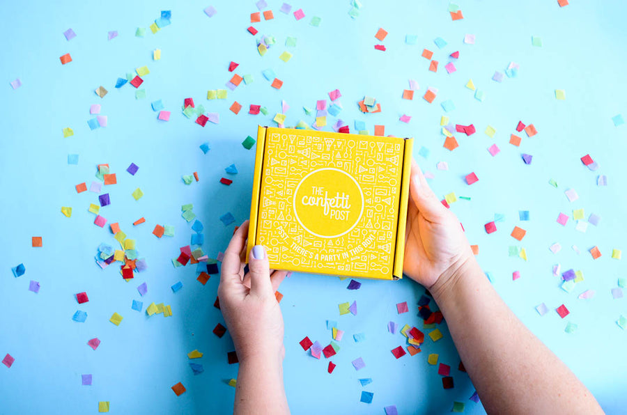 Yellow Confetti Post Gift box show closed held by two hands against a blue background with rainbow confetti scattered about